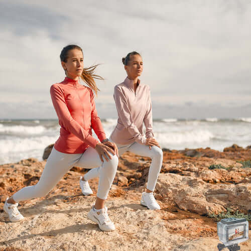 Two women in activewear demonstrating lunges on a rocky beach with ocean waves in the background.