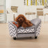 Small dog sitting on a gray and white chevron patterned pet bed in a room with chairs and a table.