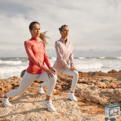 Two women in activewear demonstrating lunges on a rocky beach with ocean waves in the background.
